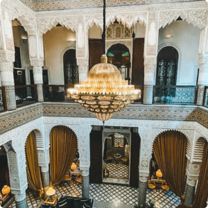 Elegant Moroccan riad interior with ornate zellige tilework, carved arches, and a grand brass chandelier – ideal for luxury travel advisors and tour operators seeking authentic, high-end accommodation options.