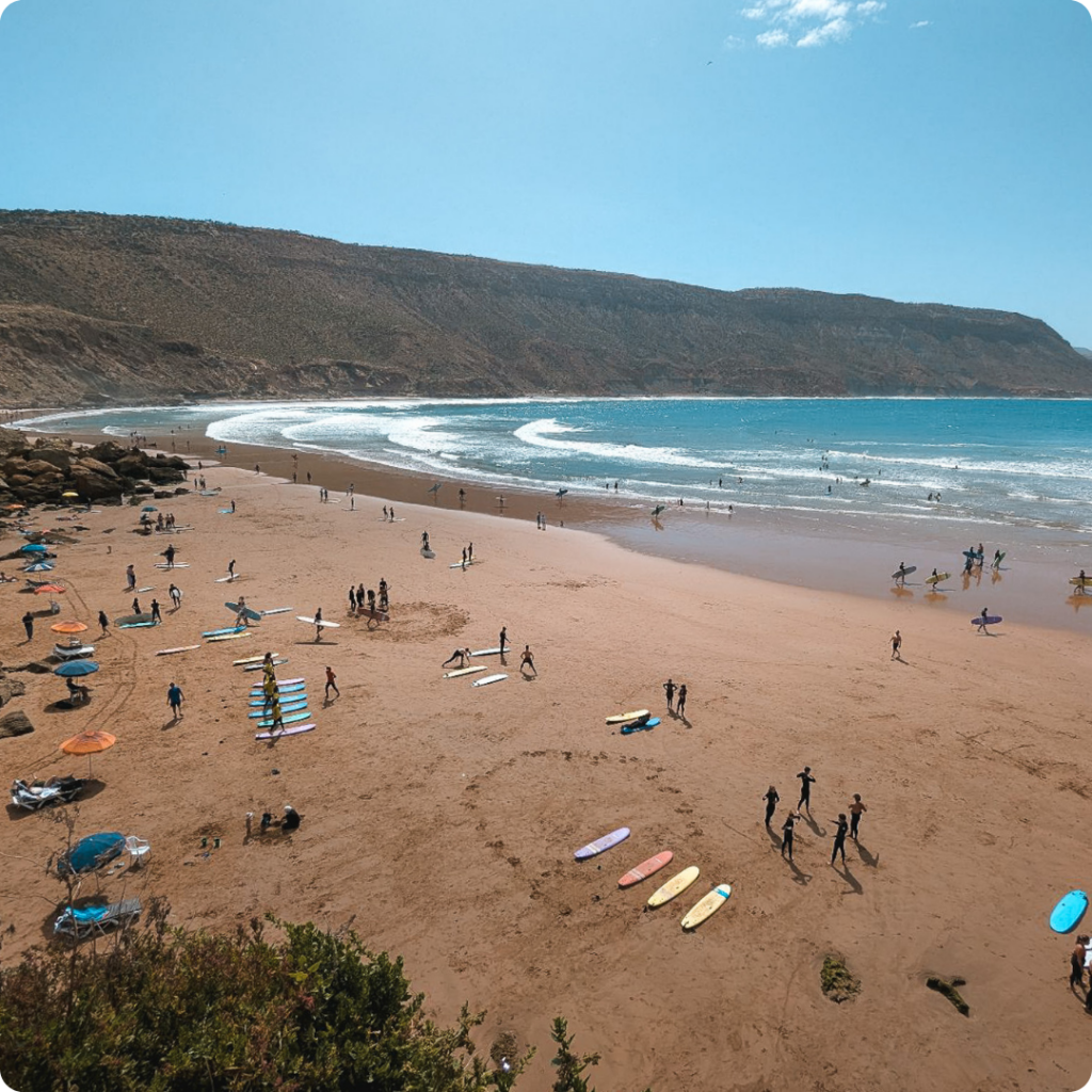 Surfers and sunseekers enjoying the waves at Morocco’s Taghazout Beach, a rising coastal hotspot for wellness retreats and surf escapes — perfect for travel advisors designing active, off-the-radar coastal itineraries.