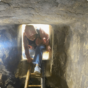 Person descending a narrow wooden ladder into the Ky Anh Tunnels in Quang Nam, Vietnam, wearing a hooded jacket and backpack, highlighting the tunnel’s small, hand-dug stone passageways.